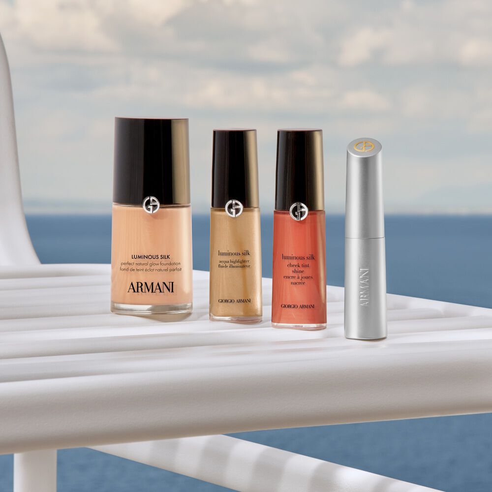 An elegant selection of Armani beauty essentials for a radiant summer glow. Featuring the iconic Luminous Silk foundation, liquid highlighter, cheek tint, and lip color, beautifully displayed against a serene Mediterranean backdrop.
