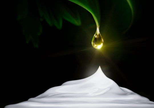 A drop of liquid gold hangs from a green leaf above a smooth, white cream or lotion, set against a dark background. A soft glow emanates from the drop.