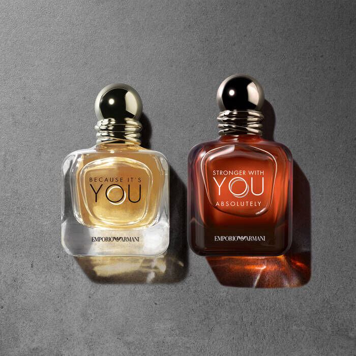 Two perfume bottles from Emporio Armani are displayed on a textured gray surface. The bottle on the left is labeled "Because It's You" with a golden liquid, while the bottle on the right is labeled "Stronger With You Absolutely" with a dark amber liquid.