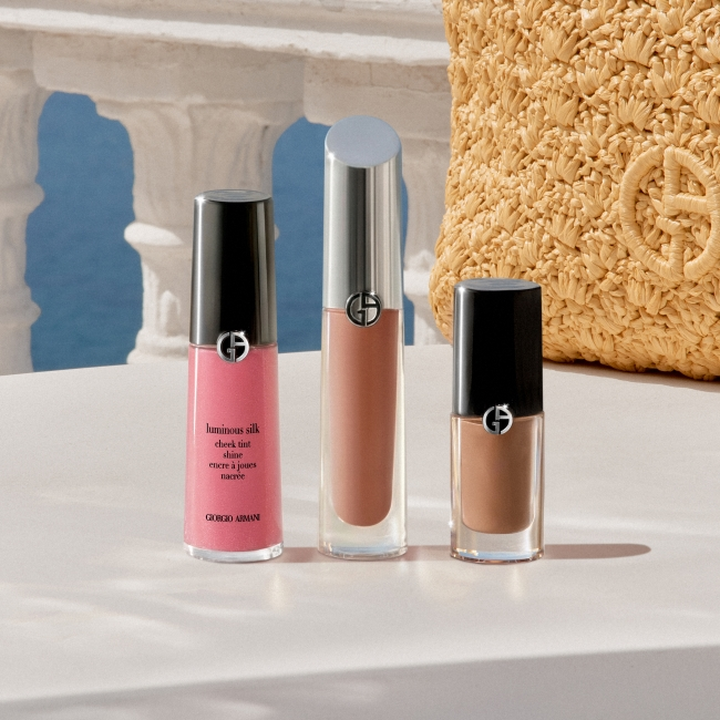 Giorgio Armani Make-up-Produkte auf einem weißen Podest: ein rosafarbenes Flussig-Rouge, ein nudefarbener Lipgloss und ein brauner Flussig-Lippenstift, vor einer mediterran inspirierten Kulisse mit Steinbogen, blauem Himmel und einem goldfarbenen Accessoire mit strukturierter Oberflache.