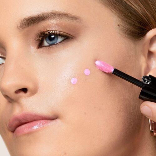 A close-up of a person's face applying pink liquid makeup with a wand, showcasing clear skin and blue eyes.