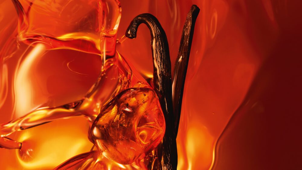 Eine Nahaufnahme von zwei dunklen Vanilleschoten und glanzendem Amberharz vor einem warmen, leuchtenden Amberhintergrund.