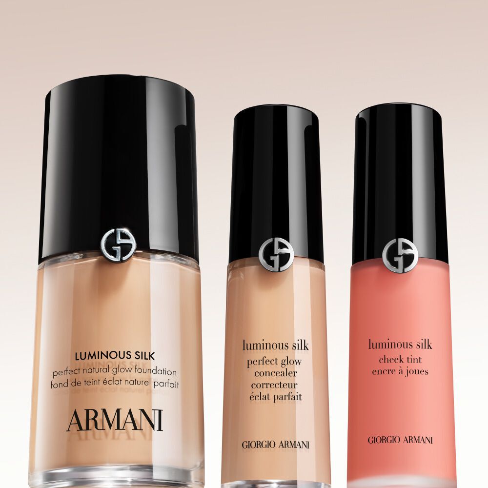 Three Armani Luminous Silk products lined up against a light background: Perfect Glow Flawless Foundation, Perfect Glow Concealer, and a coral Cheek Tint, arranged side-by-side from tallest to shortest.