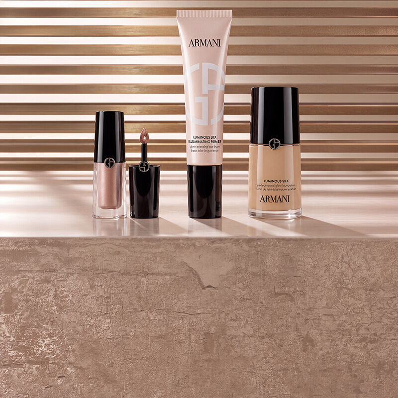 Armani beauty Make-up-Produkte, darunter Liquid Highlighter, Luminous Silk Illuminating Primer und Luminous Silk Foundation, prasentiert auf einer Marmoroberflache vor einem warmen Lamellenhintergrund.