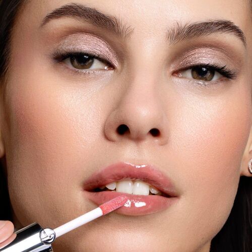 A close-up of a woman applying a glossy pink lip gloss, showcasing her well-defined makeup, including neutral eyeshadow and a radiant complexion.