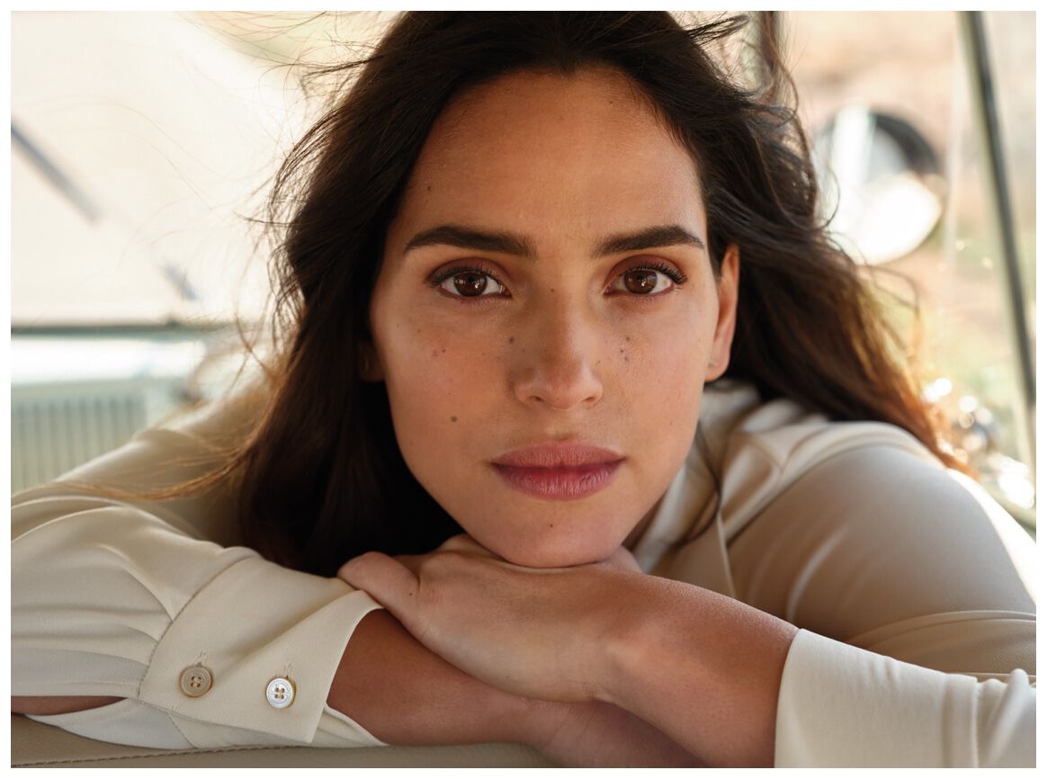 A close-up portrait of a woman with long, dark hair, resting her arms on a surface, looking directly at the camera with a thoughtful expression. She has a natural makeup look and visible freckles.