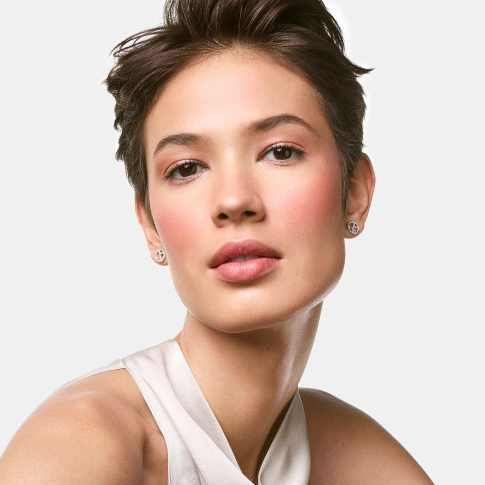 A young woman with short hair and a subtle makeup look poses confidently, wearing a simple light-colored top. She has small earrings and a neutral expression, with soft lighting enhancing her features.