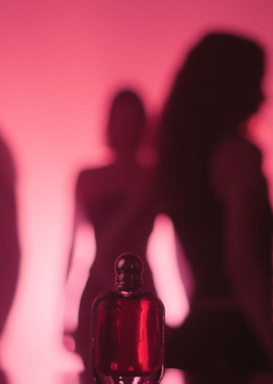 Eine stimmungsvolle, kunstlerische Anzeige fur „Power of You“ von Emporio Armani. Im Vordergrund ist der rubinfarbene Parfumflakon sichtbar, liegt jedoch teilweise im Schatten. Dahinter ist die dunkle Silhouette einer Frau, vermutlich Kendall Jenner, vor einem weichen, leuchtenden rosa-magentafarbenen Hintergrund zentriert. Weitere schwache, verschwommene Silhouetten erscheinen an den Seiten und erzeugen ein Gefuhl von Tiefe und Geheimnis. Ein Play-Button und ein Navigationspfeil sind sichtbar, was darauf hindeutet, dass dies Teil eines Videos oder einer digitalen Galerie ist.