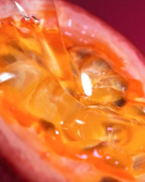 Eine extreme Nahaufnahme einer aufgeschnittenen Maracuja (Passionsfrucht), die die leuchtend orangefarbenen, saftigen Kerne und das Fruchtfleisch detailreich zeigt. Das Bild hat eine warme, glanzende Textur. In weißem Text daruber steht die Beschreibung: „Eine herb-saftige Explosion von Maracuja (Passionsfrucht) mit dem sinnlich-warmen Genuss von Vanille“. Unten befinden sich ein „ENTDECKEN“-Link und ein weißes Play-Symbol.