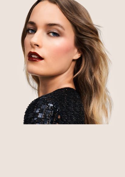 Portrat einer Frau mit langen, lockigen Haaren und einem eleganten Make-up, die seitlich in die Kamera blickt. Der Hintergrund ist neutral gehalten.