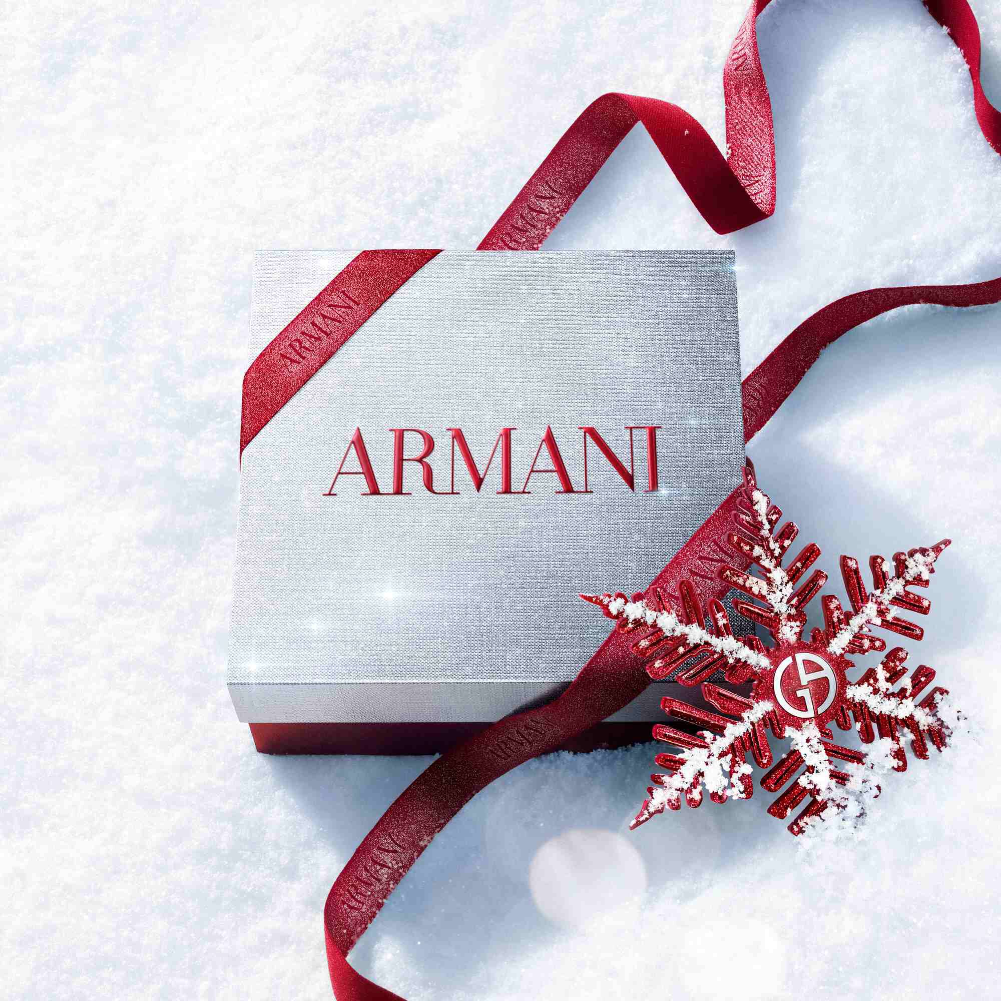 Silver Armani Glamourous Winter Holiday Gift Set box with red glitter ribbon and Giorgio Armani red snowflake ornament resting on a sparkling snowy surface — festive luxury fragrance gift packaging.