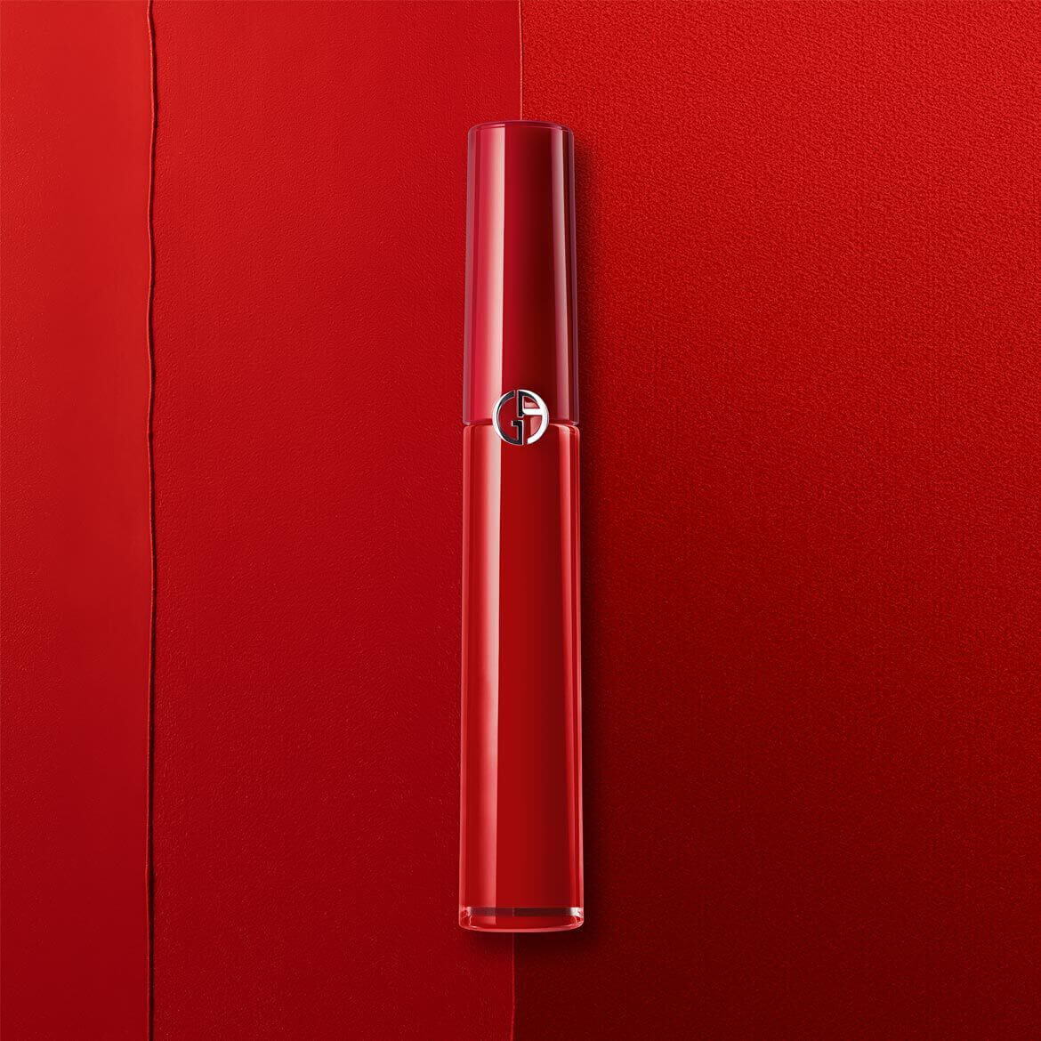 A glossy red lip product lies diagonally across a vibrant red background, showcasing a sleek design with a logo on the cap.