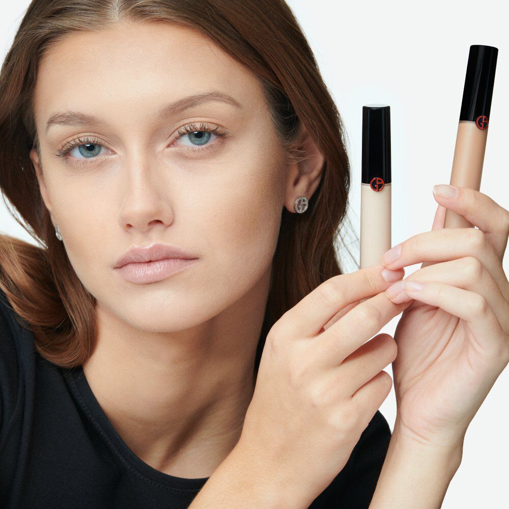 A model with long, wavy hair holds two tubes of concealer, demonstrating a natural makeup look. She has a neutral expression and is wearing a simple black top.