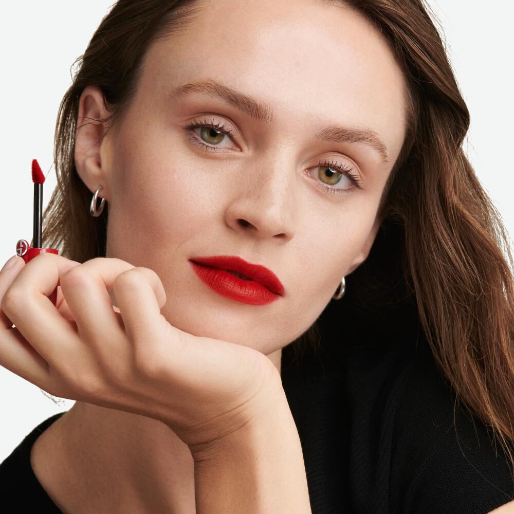A woman with long, wavy hair and striking green eyes applies vibrant red lipstick while resting her chin on her hand. She wears small silver hoop earrings and a black top.