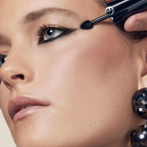 A close-up of a woman’s face with striking makeup, featuring bold eyeliner and full lashes. She appears to be applying mascara, with large, stylish earrings visible.