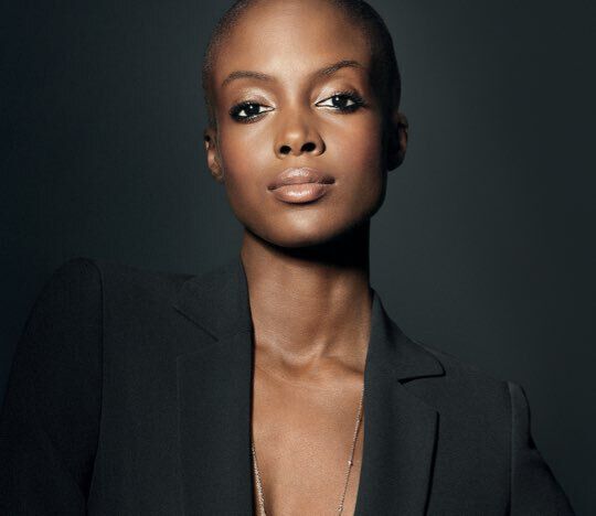 A close-up portrait of a confident person with a shaved head, wearing a dark blazer. The individual has defined facial features and is gazing directly at the camera, with a subtle, elegant makeup look.