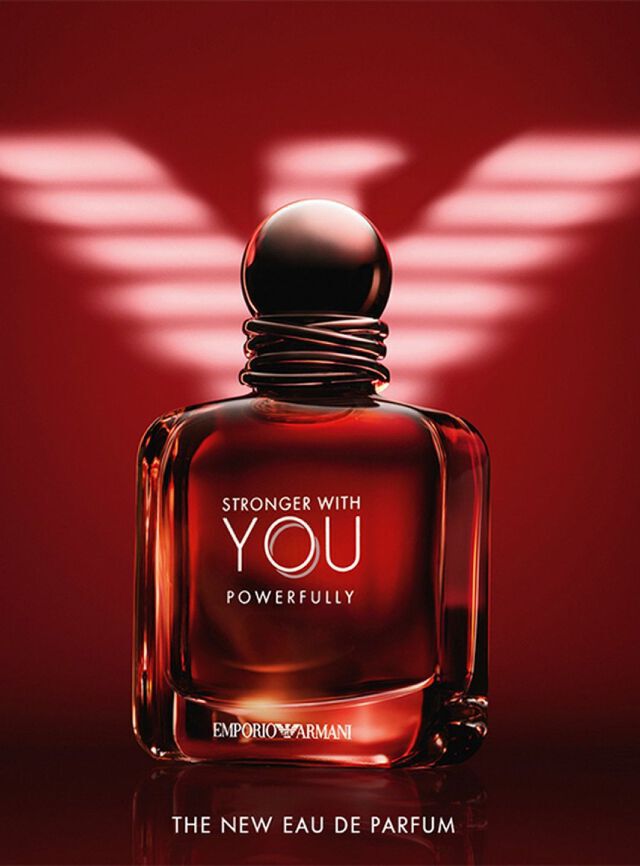 Eine dramatische Anzeige fur das „Stronger With You Powerfully“ Eau de Parfum von Emporio Armani. Das Bild zeigt einen massiven, quadratischen Glasflakon, der mit einer satten, bernsteinroten Flussigkeit gefullt ist, die von innen heraus leuchtet. Der Flakon hat einen dunklen, abgerundeten Verschluss mit einer drahtahnlichen Struktur am Flaschenhals. Die Aufschriften „STRONGER WITH YOU POWERFULLY“ und „EMPORIO ARMANI“ sind in Silber und Weiß gedruckt. Im Hintergrund ist ein großes, stilisiertes Armani-Adler-Logo in Weiß vor einem tiefroten Hintergrund beleuchtet. Unten steht der Text „THE NEW EAU DE PARFUM“.