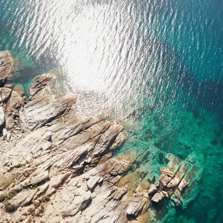 Aerial view of rocky coastline meeting clear turquoise water, with sunlight reflecting off the surface.