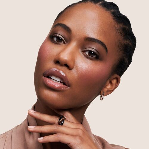 Portrat einer Frau mit geflochtenem Haar, leichten Wangenrouge und modischem Schmuck, vor neutralem Hintergrund.