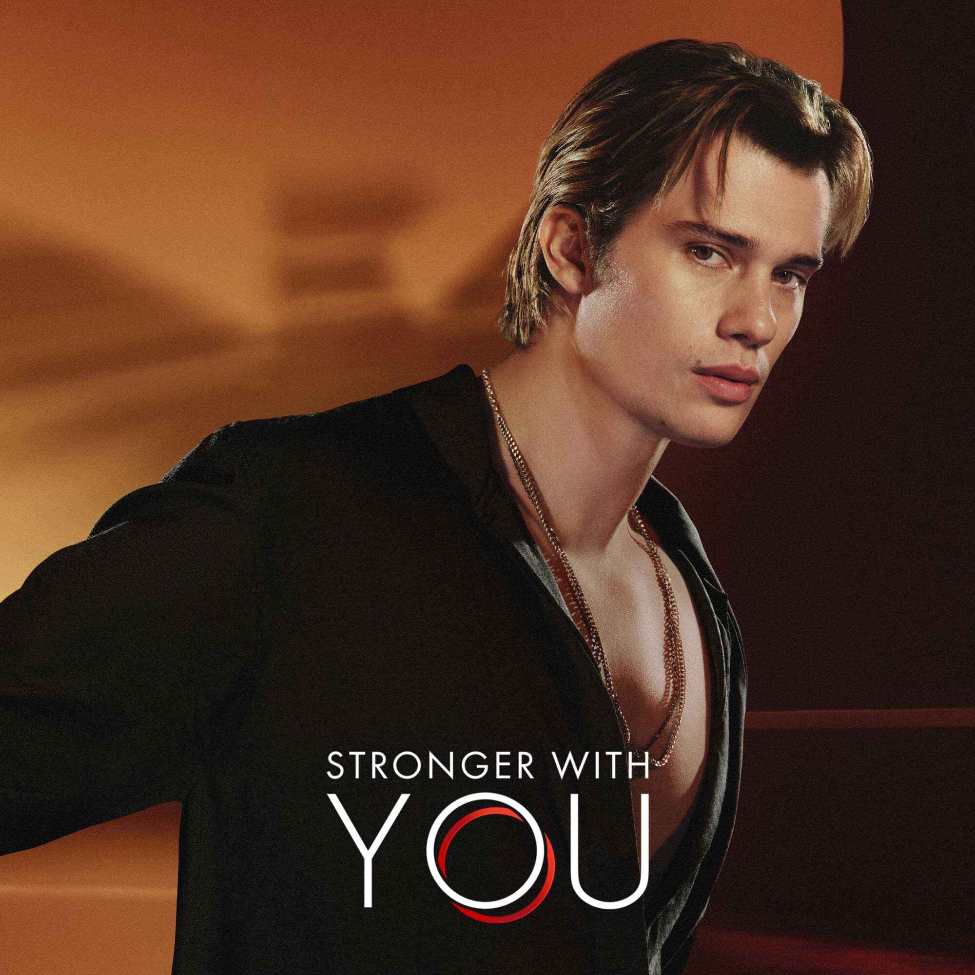 Nicholas Galitzine in einer stilvollen Armani-Kampagne fur den Duft „Stronger With You“, in einem schwarzen, offenen Hemd vor einem warmen, eleganten Hintergrund.