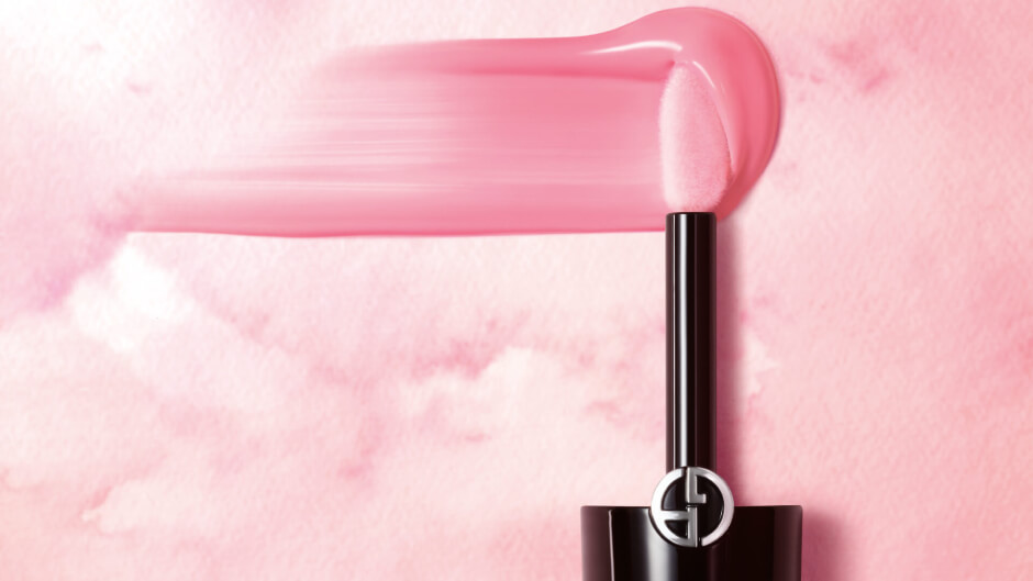 A sleek lipstick applicator with a vibrant pink gloss, set against a soft pink watercolor backdrop.